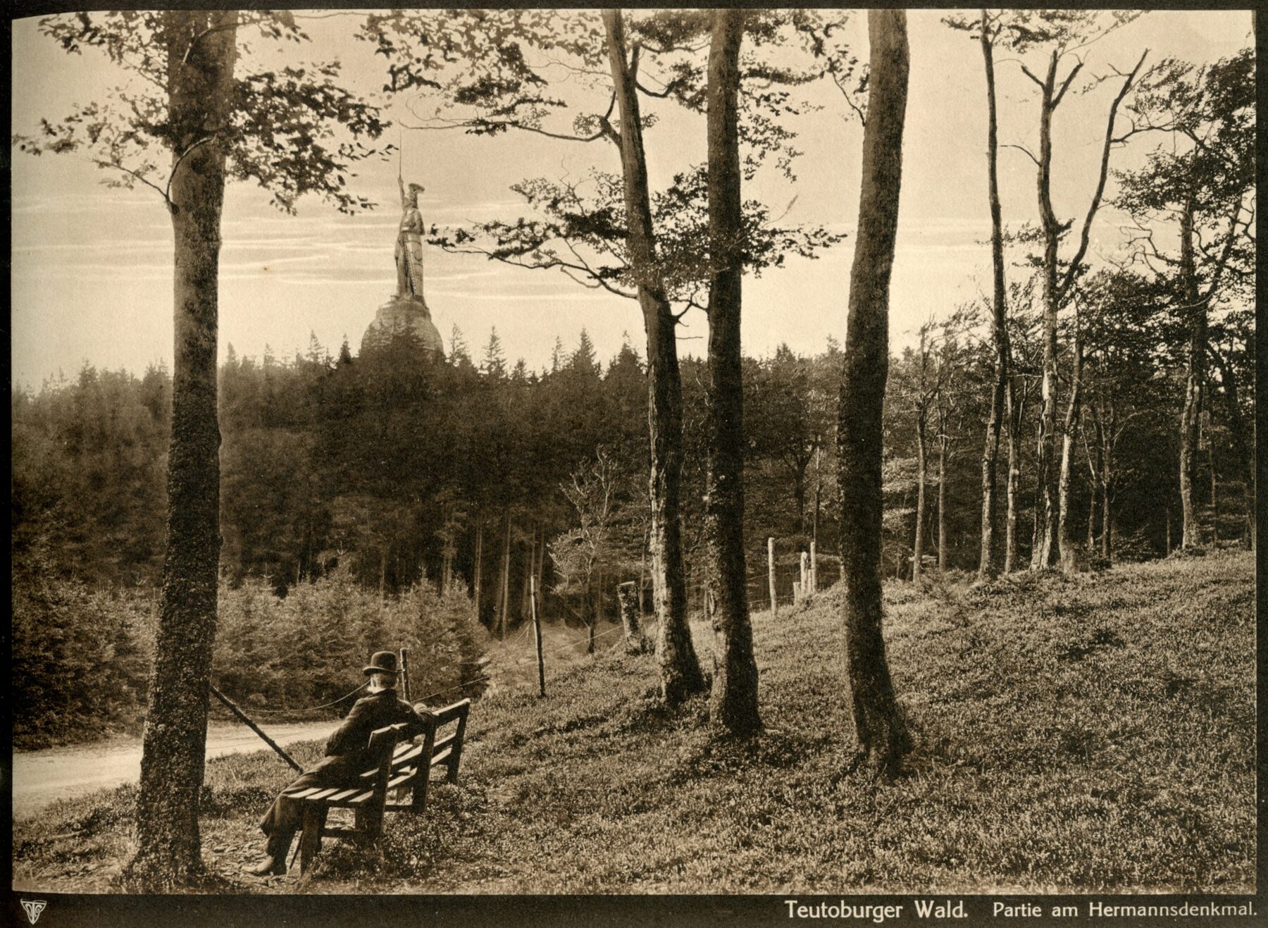 Blick auf das Hermannsdenkmal von einem Wanderweg mit Bank, auf der ein Spaziergänger sitzt